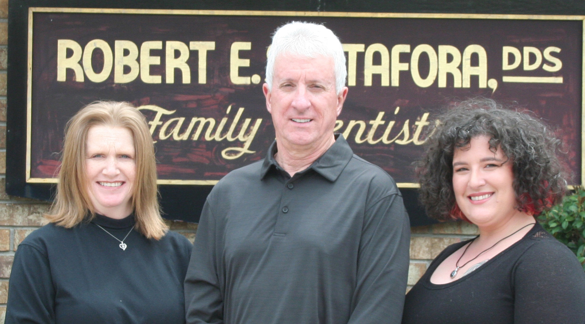 The image shows a group of three adults posing in front of a sign for  Robert E. Sapora DDS Family Dentist.  They are standing outdoors, with the person on the left wearing a black shirt and the other two dressed in dark tops.