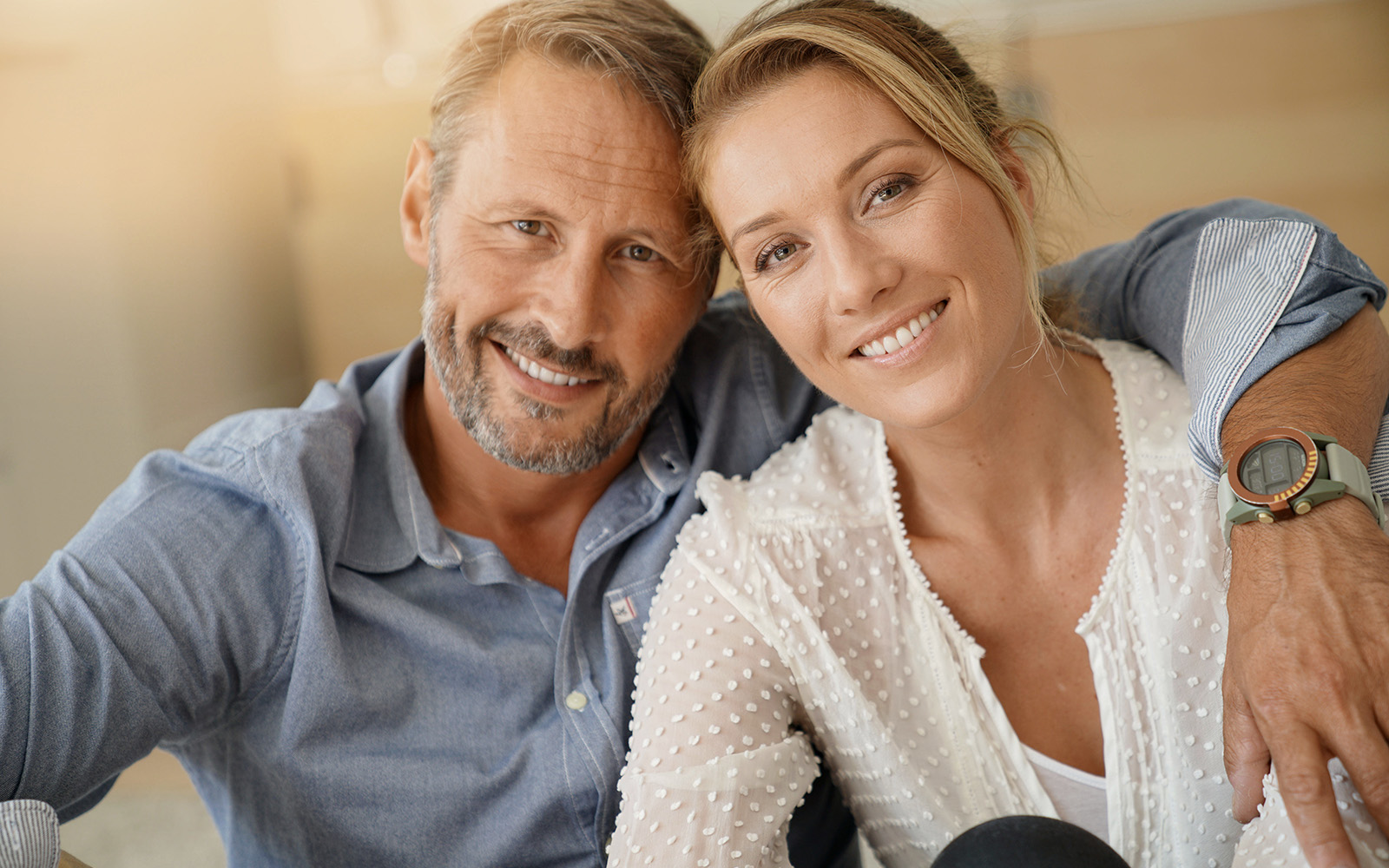 The image is a photograph of an older man and woman posing together, with the man standing behind the seated woman, both smiling at the camera.