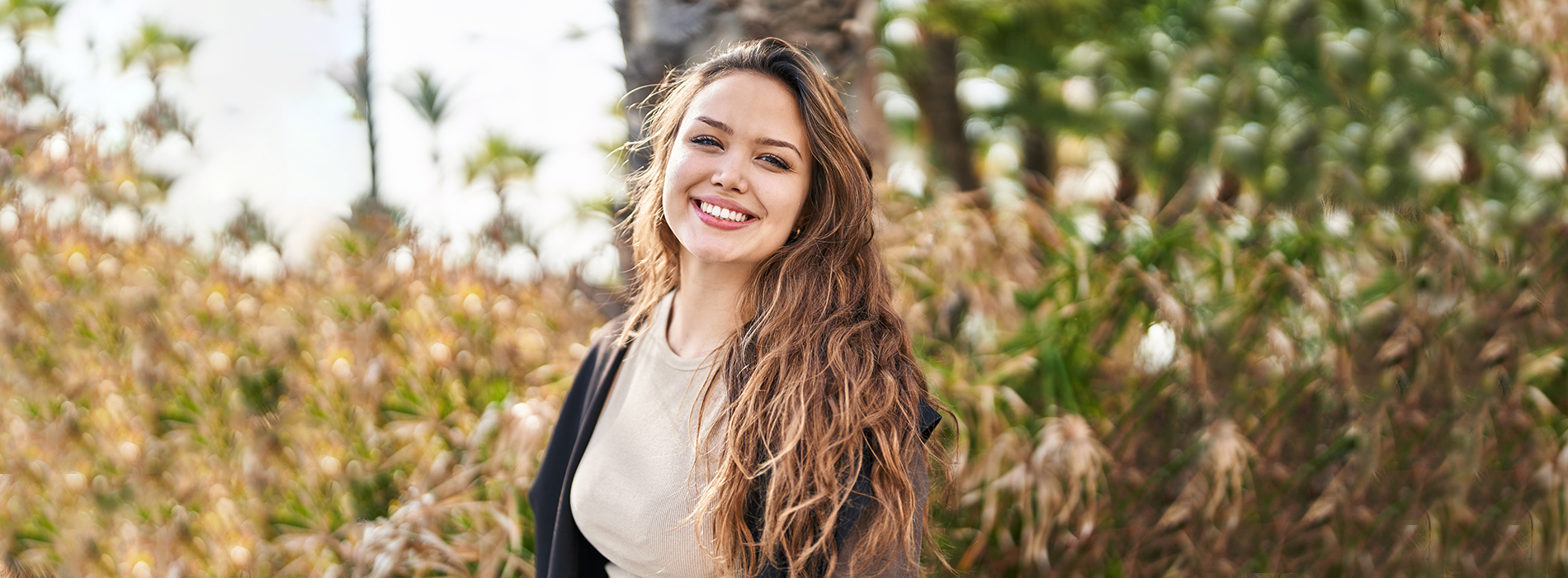 A woman with long hair, smiling and standing in front of a hedge.