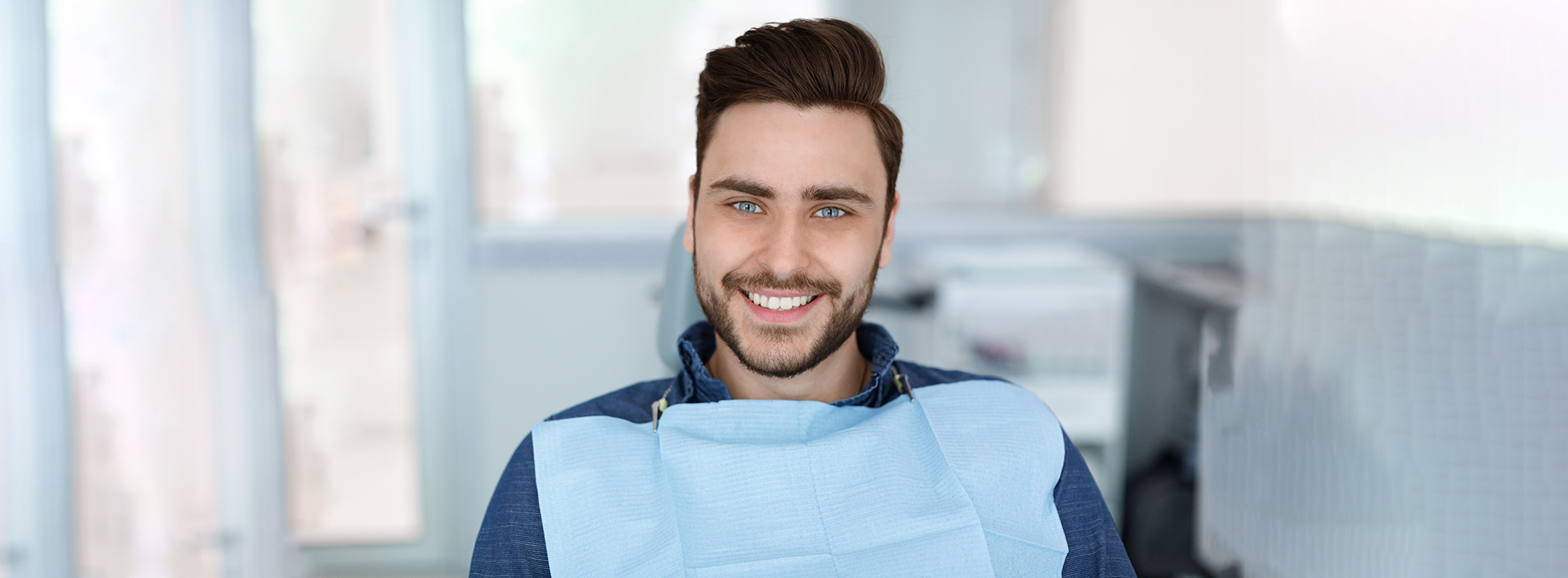 A man in a dental office, smiling at the camera while wearing a blue shirt and sitting in front of a desk.