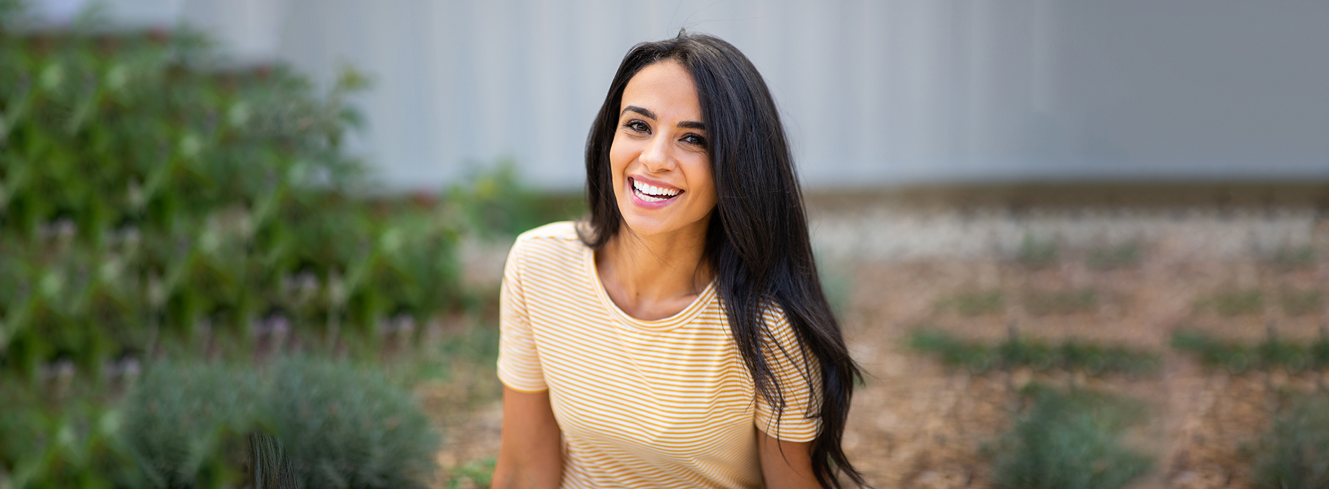 The image is a photograph of a smiling woman with long hair, wearing a yellow top, standing outdoors in front of a building and plants.