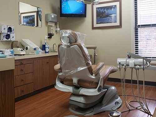 An image of a dental office interior, featuring a dental chair with plastic wrap on it and various dental equipment in the background.