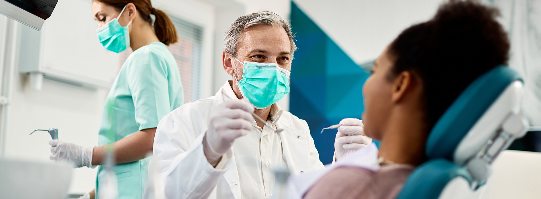 Dental professionals in a clinic setting, one person seated and smiling at the camera with gloved hands, another standing behind, both wearing masks and surrounded by dental equipment.