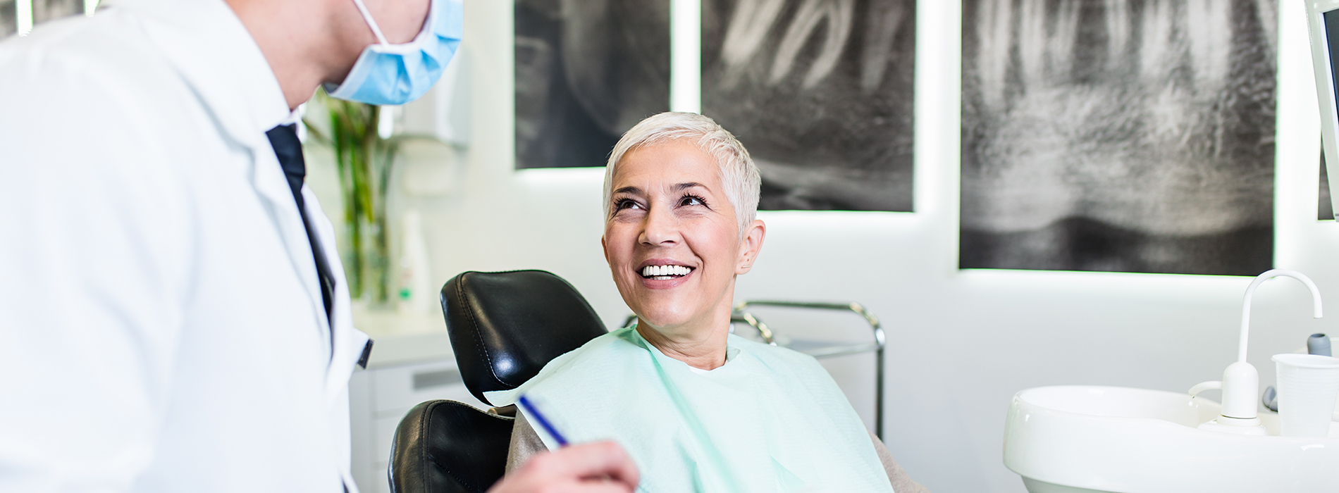 A dentist in a modern dental office, smiling and looking at the camera while a patient is seated in the chair.