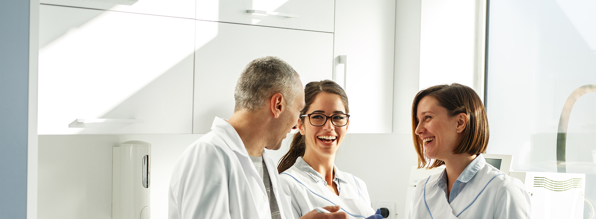 The image shows a group of three people, two women and one man, in a professional setting, possibly an office or laboratory, wearing white lab coats, engaged in a conversation.