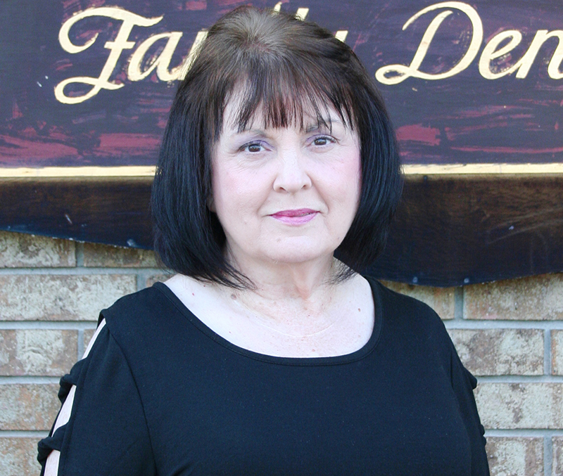 The image is a photograph of a woman standing in front of a sign that reads  Family Dental.  She has short dark hair and is wearing a black top.