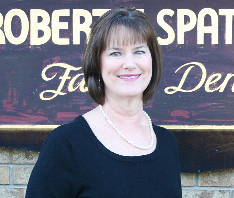 The image shows a woman standing in front of a sign that reads  Robert Spat.  She is wearing a black top and has short hair.