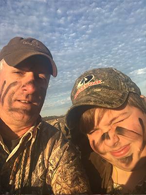 The image shows a man and a young boy, both wearing camouflage clothing and hats, standing together outdoors. They appear to be engaged in an activity related to hunting or the outdoors, possibly posing for a photograph.
