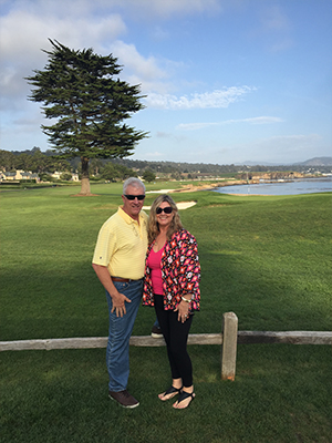 A man and a woman are standing on a golf course, posing for a photo in front of a scenic view.