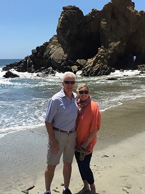 An elderly couple standing on a sandy beach, posing for a photo in front of the ocean with rock formations and clear skies.