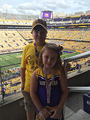 An image of two individuals, an adult and a child, seated in the stands at a football stadium during a game, with the adult wearing a yellow jersey and the child in a purple one.