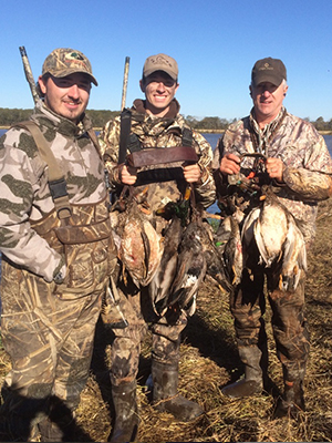 Three men posing with ducks in a natural setting, likely hunters showing off their catch.