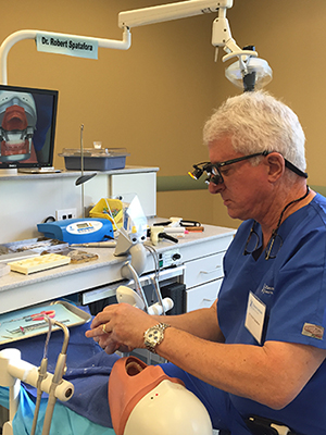 An elderly man in a dental office, wearing glasses and working on a dental chair with various tools around him.
