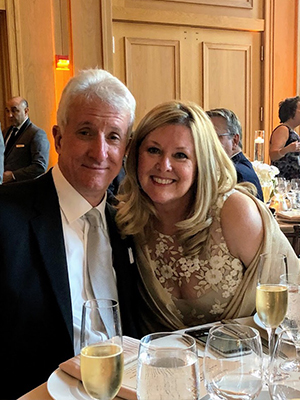 A man and a woman posing at a formal event, seated at a dining table with wine glasses and a menu in front of them.