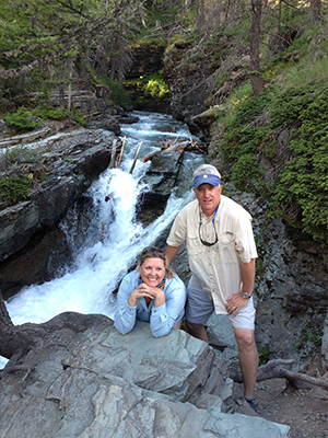The image shows a man and woman posing in front of a scenic waterfall with a stream flowing through it, surrounded by lush greenery.