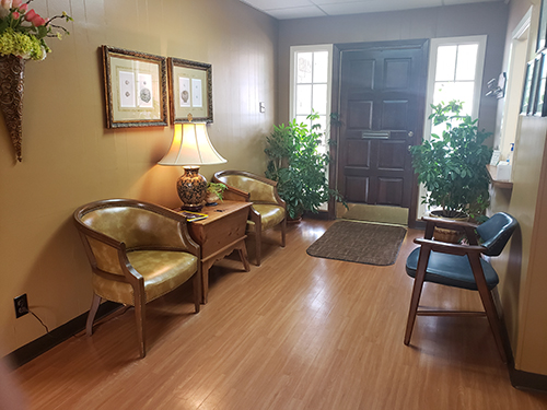 The image depicts an interior space, likely a waiting or reception area, with furniture including chairs and a couch, a framed picture on the wall, a potted plant, and a door leading to another room.