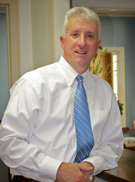 The image shows a man wearing a white shirt and blue tie, standing in an office setting.