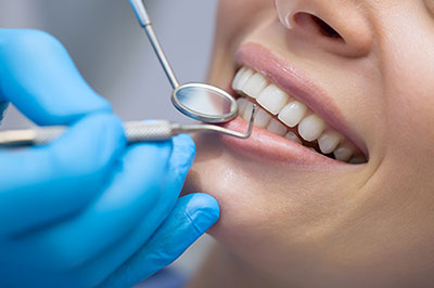 The image shows a dental professional performing oral care on a patient, with the patient smiling while wearing a surgical mask and having their teeth cleaned.