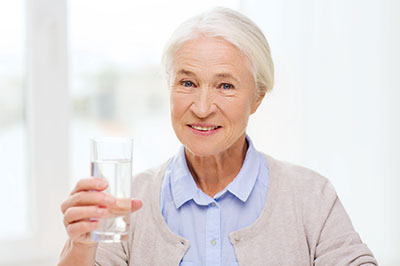 An elderly woman is holding a glass of water to her lips, smiling.