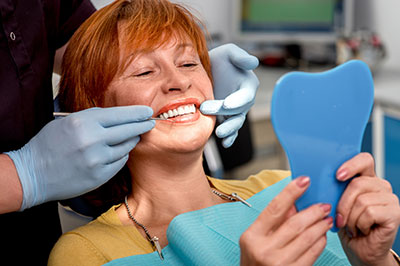 The image shows a woman sitting in a dental chair, smiling at the camera while holding a blue dental impression tray. She has a toothpick in her mouth, and there is a dental professional standing behind her, wearing gloves and a surgical mask, examining the tray with a smile.