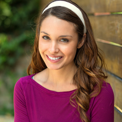 A woman with long brown hair and a headband is smiling at the camera. She has a purple top on and appears to be outdoors near a wooden fence.