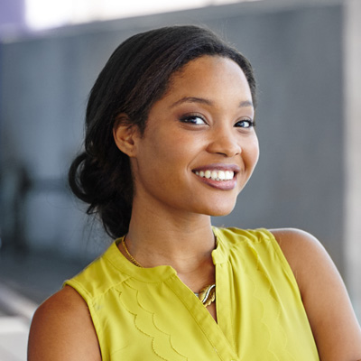 This is a photograph of a smiling woman with dark hair, wearing a yellow top and standing in front of a building.