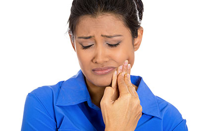 A woman with dark hair, wearing a blue blouse and a white mask, appears concerned as she looks at her hand and mouth.