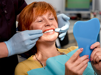 A woman in a dental chair receiving a teeth cleaning, with a dentist holding a blue model of a mouth and smiling.