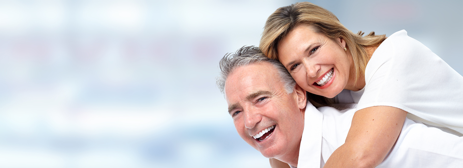 The image is a photograph of two older adults, a man and a woman, smiling and embracing each other. They appear to be happy and are dressed in casual attire. The background is blurred but suggests an indoor setting with warm lighting.