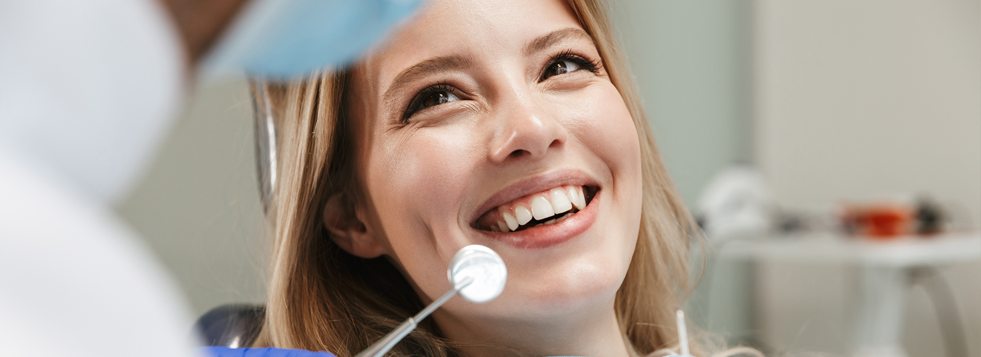 A woman with a smile is sitting in front of a dental professional, who appears to be examining her teeth.