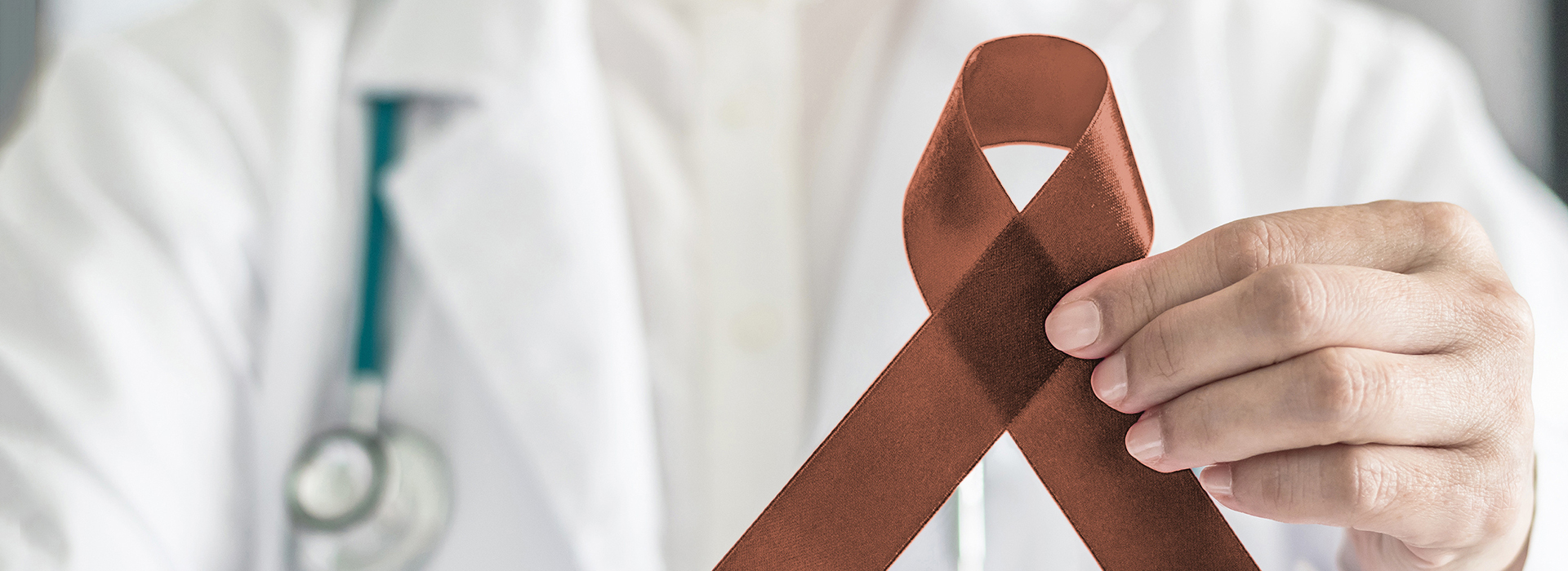 The image shows a medical professional holding a brown ribbon with a red symbol in their hand, against a backdrop of a white lab coat.