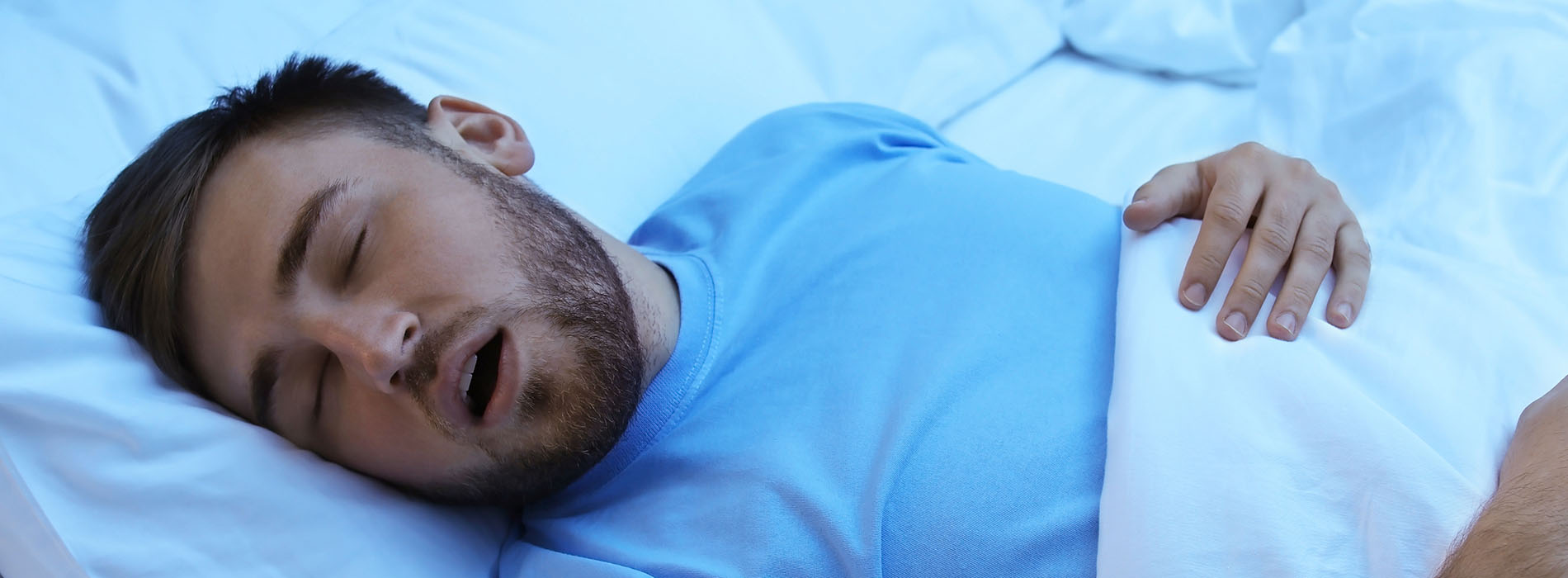 A man lying in bed, with his eyes closed and mouth open, suggesting he is asleep or possibly snoring.