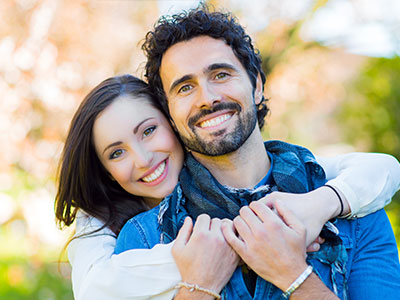 A man and a woman are smiling at the camera, with the man in the foreground and the woman slightly behind him. They appear to be a happy couple.