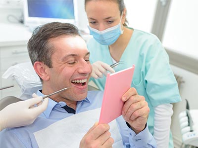 A man is seated in a dental chair, holding a pink card with both hands, and smiling at the camera. Behind him, a female dental professional is standing, looking at the man s card with a stethoscope around her neck.