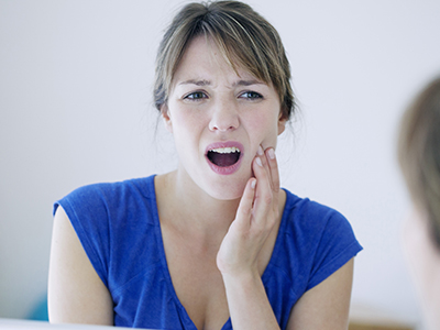 The image depicts a woman with a concerned expression, looking at her reflection in the mirror while holding her hand near her mouth.