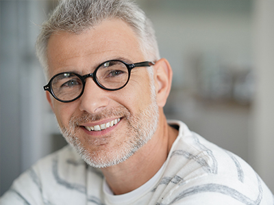 The image is a portrait of a man with glasses, smiling at the camera. He has short hair and appears to be middle-aged. The background is blurred but suggests an indoor setting, possibly a home or office environment.