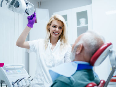 A dental hygienist in a white uniform is assisting an older man with a blue mask, who is seated in a dentist s chair.