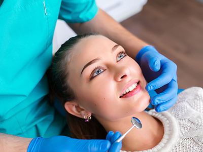 The image depicts a dental professional performing an examination on a patient s teeth, with the patient seated in a chair and wearing glasses.