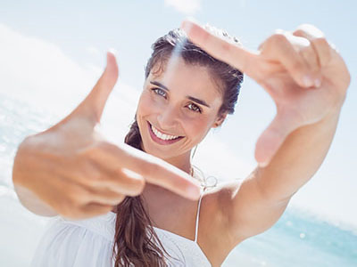 A woman is taking a selfie with both hands, smiling and looking at the camera. She has long hair, is wearing a white top, and is standing by the beach with clear skies in the background.